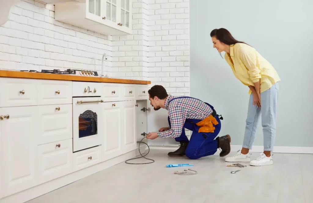 Top Sump Pumps. Plumber crouching on the floor in a modern white kitchen and using a drain cable to clean a clogged sink pipe